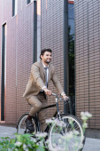 šťastný muž v obleku jízda na kole a usmívání v blízkosti budovy a rostlina na rozmazané popředí 