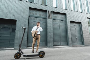 Takım elbiseli bir işadamı e-scooter ve modern binanın yanında dikilirken akıllı telefondan konuşuyor.