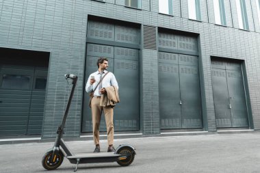 E- scooter ve binanın yanında dururken akıllı telefon tutan işadamlarının tam uzunluğu 