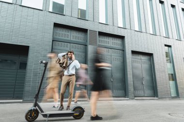 Takım elbiseli bir işadamı e-scooter ve insanların yanında dururken akıllı telefondan konuşuyor, hareket bulanıklığı
