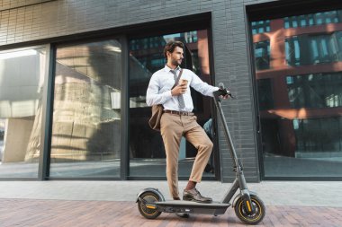 Elektronik scooter kullanan bir işadamı elinde kağıt bardakla modern caddede