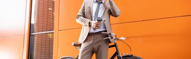 partial view of businessman holding paper cup near bicycle and building with orange walls, banner clipart