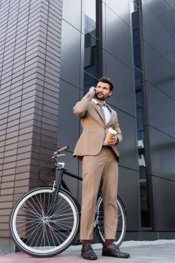 Düşük açılı iş adamı görüşü akıllı telefonla konuşuyor ve bisiklet ve binanın yanında kağıt bardak tutuyor. 