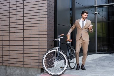 full length of smiling businessman holding smartphone and coffee to go near bicycle while standing near building  clipart