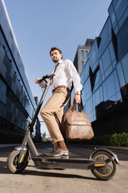 Resmi olarak sakallı iş adamının dar açılı görüntüsü deri çanta taşıması ve elektrikli scooterın yanında durması. 