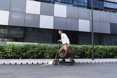 Resmi kıyafetli sakallı iş adamı elektrikli scooter kullanıyor ve elinde çanta tutuyor.