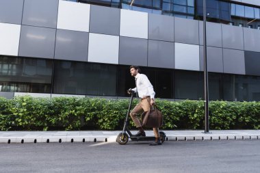 Resmi kıyafetli sakallı iş adamı elektrikli scooter kullanıyor ve elinde çanta tutuyor.