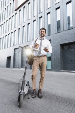 Kağıt bardak tutan ve e-scooter 'ın yanında akıllı telefon kullanan takım elbiseli neşeli bir işadamı. 