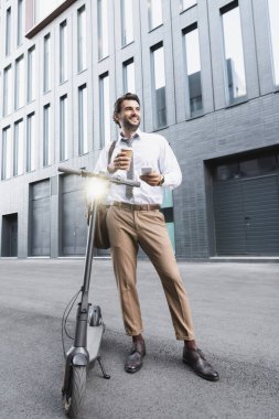 Resmi olarak neşeli iş adamları ellerinde kağıt bardakla e-scooter 'ın yanında akıllı telefon kullanıyor. 