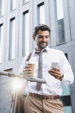 Resmi olarak gülümseyen işadamı elinde kağıt bardakla e-scooter 'ın yanında akıllı telefon kullanıyor. 