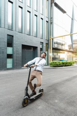 amazed businessman in formal wear riding electric scooter near building  clipart