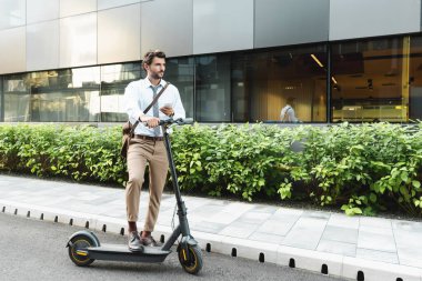 E- scooter, fabrika ve binanın yanında dururken akıllı telefon tutan iş adamlarının tam uzunluğu 