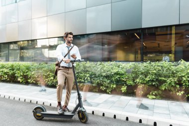 Akıllı telefon tutan iş adamının hareket bulanıklığı e-scooter, fabrikalar ve binaların yanında dururken 