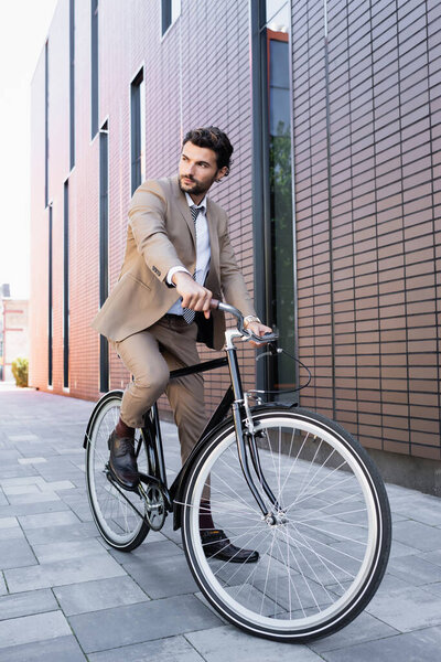 full length of bearded man in suit riding bicycle and looking away near building 
