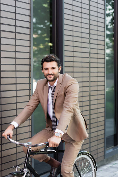 happy businessman in suit riding bicycle and looking at camera near building 