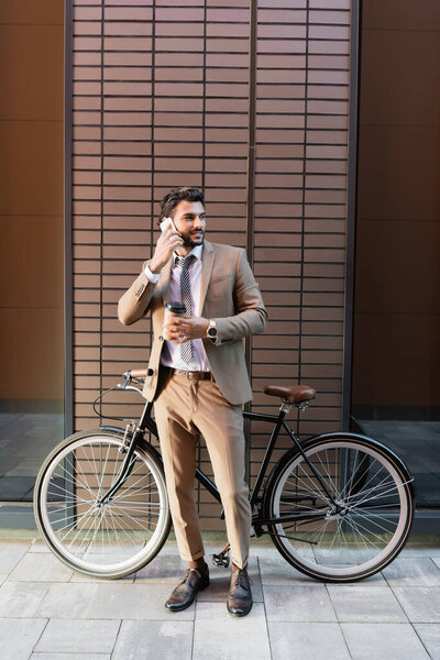 full length of bearded businessman talking on smartphone and holding paper cup near bicycle and building 