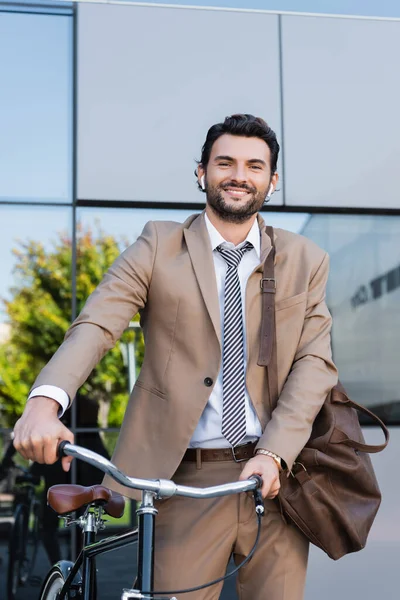 smiling businessman in wireless earphones while standing with bag near bike and building