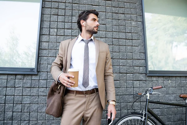 businessman in wireless earphones and suit holding paper cup and smartphone near building