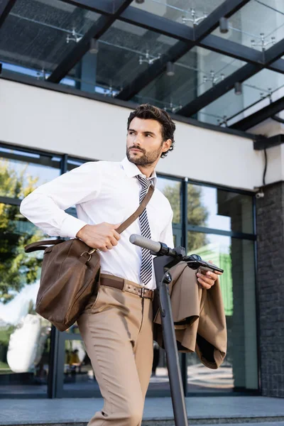 businessman in formal wear with leather bag standing near e-scooter and holding jacket