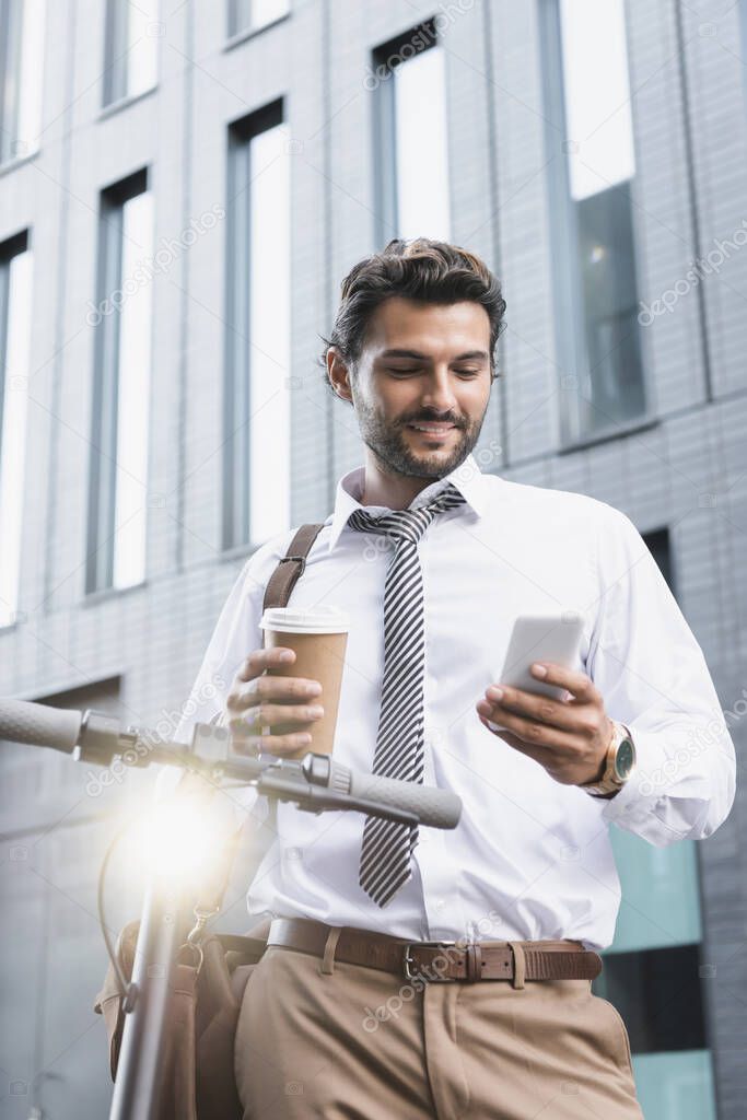 smiling businessman in formal wear holding paper cup and using smartphone near e-scooter 