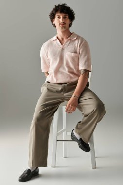 Kendine güvenen genç adam zarif bir şekilde bir taburede oturuyor, şık giyinmiş ve karizma yayıyor..