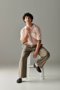 Moda kıyafetleri giymiş yakışıklı bir genç şık bir ortamda taburede dengeli bir poz verir..