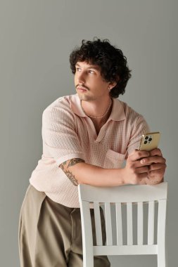 Pembe bluzlu ve bej pantolonlu genç bir adam derin düşüncelere dalmış bir telefon tutuyor..