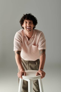 Şık bir genç adam bir tabureye yaslanıyor, elbisesini ve parlak gülümsemesini sergiliyor..