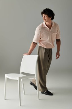 Şık bir genç adam beyaz bir sandalyenin yanında duruyor, minimalist bir alana güven yayıyor..