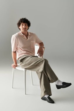 Yakışıklı genç adam kendine güvenerek bir sandalyeye oturuyor, parlak bir yerde moda seçimleri sergiliyor..