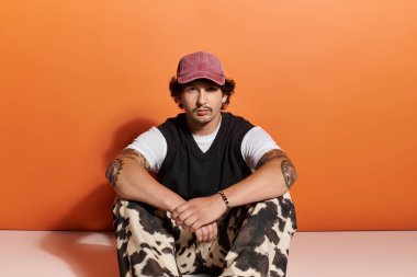 Şık giyinmiş kendine güvenen genç bir adam turuncu bir arka planda yerde oturuyor..