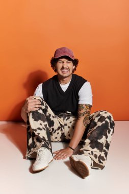 Şık bir genç adam parlak turuncu bir arka planda gelişigüzel oturuyor, kendine güveni ve cazibesi var..