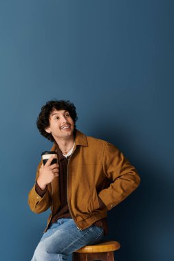 Yakışıklı genç bir adam bir taburede oturuyor, elinde bir fincan tutarken gülümsüyor, ışık saçıyor..