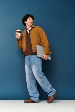 Yakışıklı genç adam elinde bir kahve fincanı ve dizüstü bilgisayarla, modaya uygun giyinmiş bir şekilde duruyor..