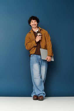 Kıvırcık saçlı şık genç adam elinde bir kupa ve dizüstü bilgisayarla mavi bir duvara yaslanmış gülümsüyor.