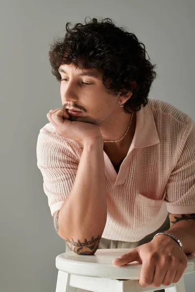 Kıvırcık saçlı genç bir adam çenesini düşüncelere dalmış modaya uygun kıyafetler giyiyor..