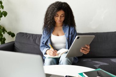Genç bir Afrikalı Amerikalı kadın bir deftere yazıyor ve evde bir tablet kullanıyor..