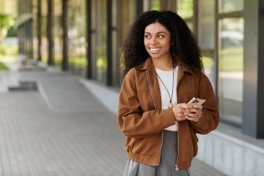 Kıvırcık saçlı genç bir kadın, güz kıyafetleri içinde telefonunu tutarken gülümsüyor..