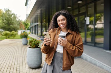 Güzel kıvırcık saçlı, sonbahar kıyafeti giymiş genç bir kadın elinde kahve tutarken gülümsüyor..