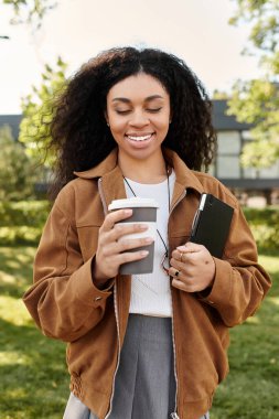 Kıvırcık saçlı genç bir Afrikalı Amerikalı kadın parkta içki içerken gülümsüyor..