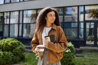 Kıvırcık saçlı genç kadın dışarıda sonbahar havasını kucaklarken elinde bir kahve ve akıllı telefon tutuyor..