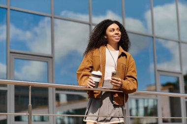 Güzel bir sonbahar gününde elinde kahve fincanı ve telefonla gülümseyen kıvırcık saçlı rahat bir kadın..