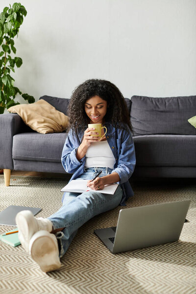 Молодая афроамериканка наслаждается работой из дома, сидя на полу с ноутбуком.