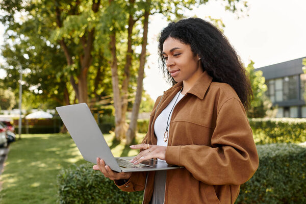 A confident Black woman in a chic autumn outfit uses her laptop in a sunny park.