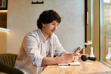 Genç bir adam ahşap bir masada rahat bir şekilde oturur, modern bir kafe ortamında akıllı telefonuna odaklanır..