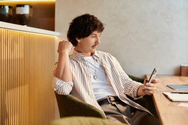 Yakışıklı genç bir adam modern bir kafede rahat bir şekilde oturuyor, akıllı telefonuyla meşgul..