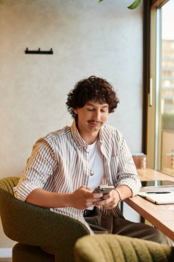 Genç ve çekici bir adam modern bir kafede rahatlar, akıllı telefonunu gülümseyerek karıştırır..