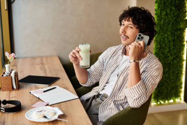 Yakışıklı genç bir adam kafede rahatlar, telefonda sohbet ederken soğuk bir içecek yudumlar..