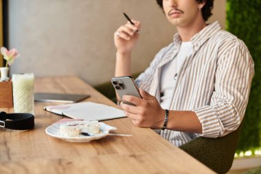 Yakışıklı genç bir adam kafeteryada oturuyor, telefonuna odaklanmış, tatlıyla çevrili..