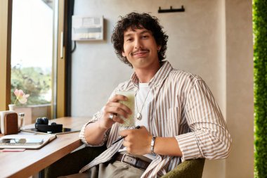 Tarz sahibi genç bir adam bir kafede rahatlar, bir içkinin tadını çıkarır ve neşeli bir şekilde gülümser..